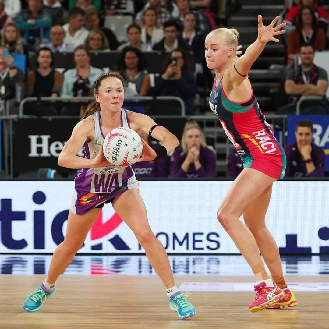 Caitlyn Strachan in the Queensland Firebirds purple Wing Attack bib holding the ball, defended by a Melbourne Vixens player at full extension.