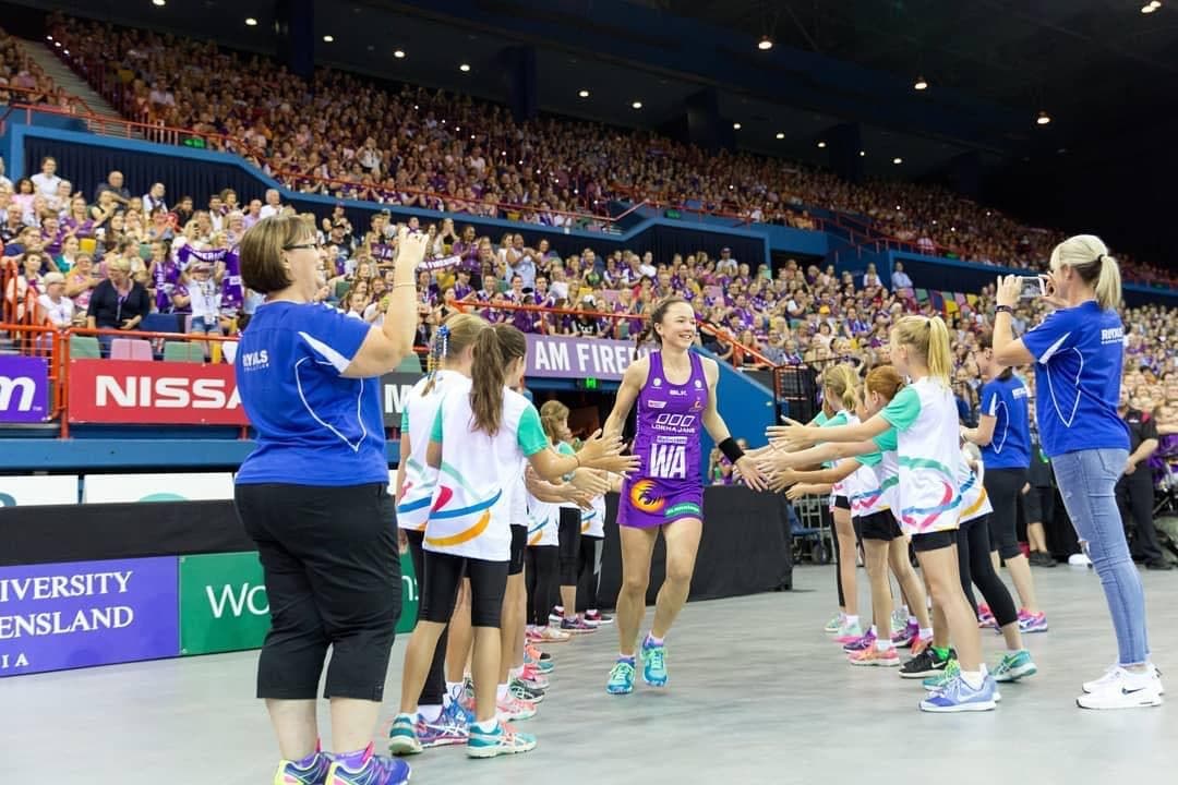 Caitlyn running onto court at a Firebirds home game, high-fiving a guard of honour of young fans in a packed Brisbane crowd.