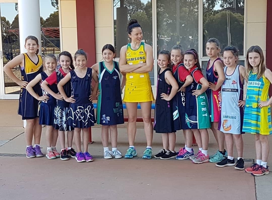 Caitlyn Strachan in the Diamonds gold standing with a line of young players in their club netball dresses, all in matching power poses.