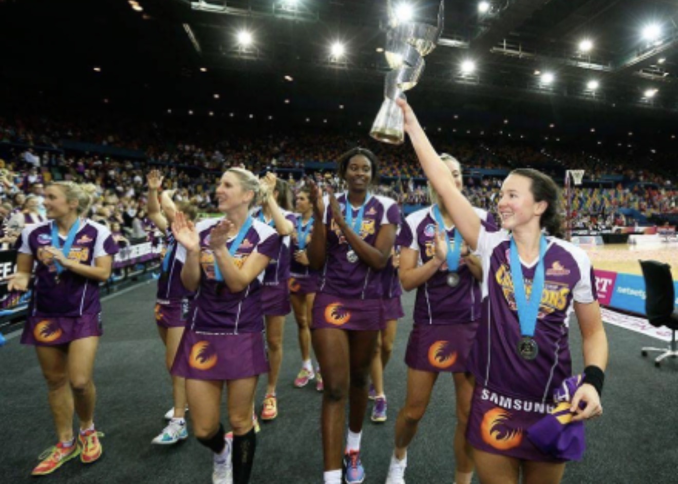 Caitlyn Strachan lifting the championship trophy on the Firebirds premiership lap of honour, wearing a Champions tee.