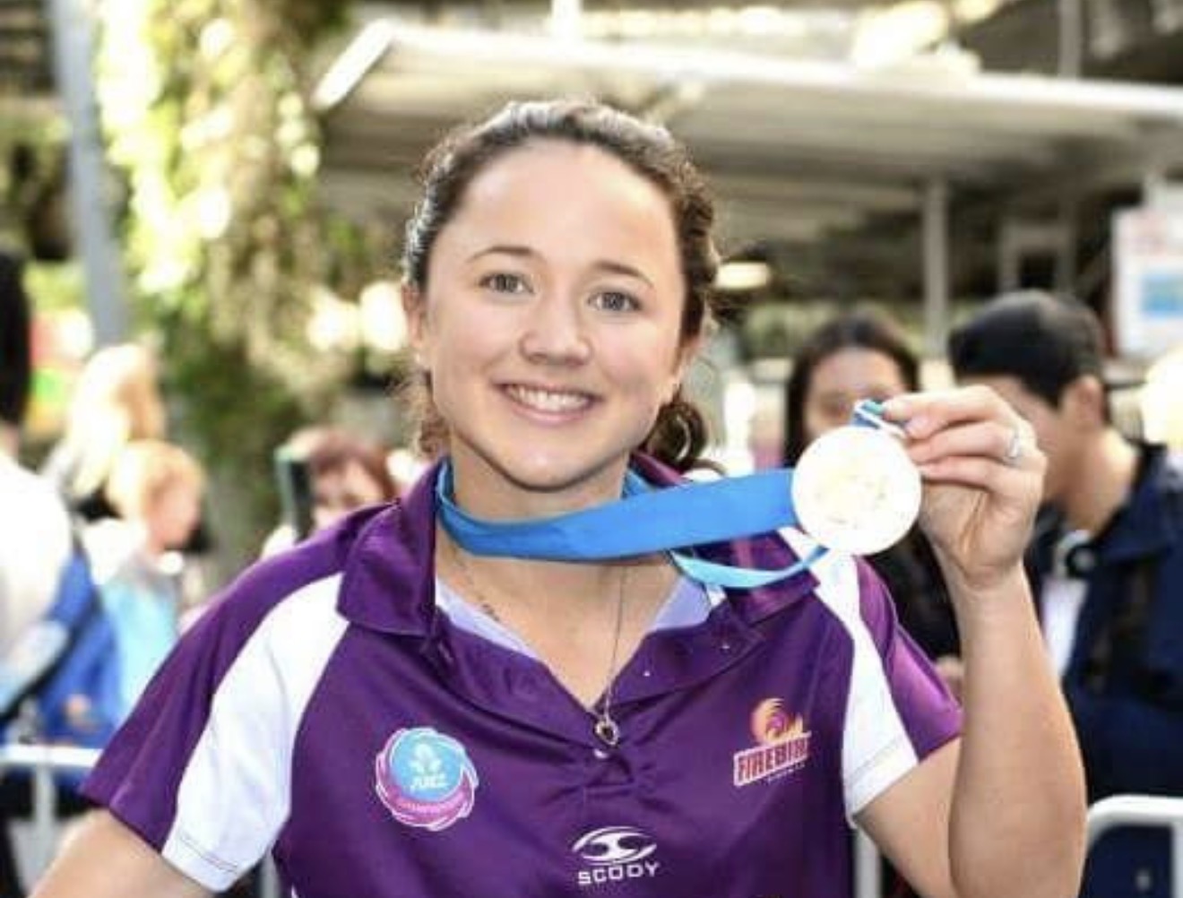Close-up portrait of Caitlyn Strachan smiling and holding up her ANZ Champions medal in a Firebirds polo.